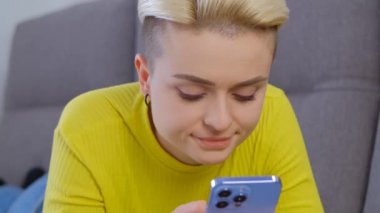 Evdeki kanepede yatarken cep telefonuna mesaj yazan genç bir kadın. Kısa boyalı saçlı beyaz kadın internet iletişimi için modern akıllı telefonu kullanıyor. Erkek Fatma çevrimiçi yorum yazıyor