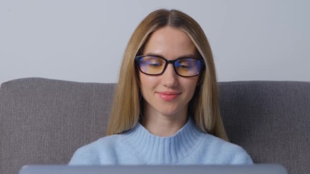 自宅のソファに座っている間 ノートパソコンで働く眼鏡の女性 若い大人のブロンドの女性は ロックダウンで遠くの仕事をオンラインでやっている 4Kストックビデオでノートパソコンを使用しているフリーランスの人 — ストック動画