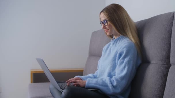 起業家の女性はロックダウンで自宅で働いています リビングルームのソファに座っている間 現代のラップトップキーボードにテキストを入力する眼鏡のブロンドの女性 遠くの仕事をするフリーランスに焦点を当て — ストック動画