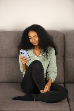 Evdeki kanepede otururken cep telefonuna göz atan kafası karışmış siyahi bir kadın. Tecritteyken modern akıllı telefon kullanan kafası karışık, mutsuz bir Afrikalı kadın. Genç yetişkin kişi sosyal medyaya göz atıyor 