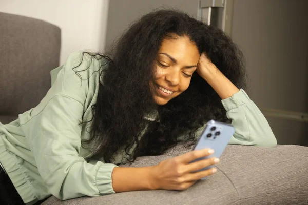 Beautiful young black woman dating online in social media app. Cheerful African female with curly hair lying on couch at home and using modern blue smartphone for entertainment and communication
