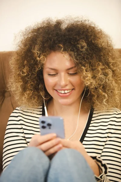 ヘッドフォンで音楽を聴いたり 携帯電話アプリでメッセージを入力したりする 巻き毛やめまいのある若い女性が興奮しています オンライン通信のための新しいスマートフォンを使用して陽気な白い女性 — ストック写真