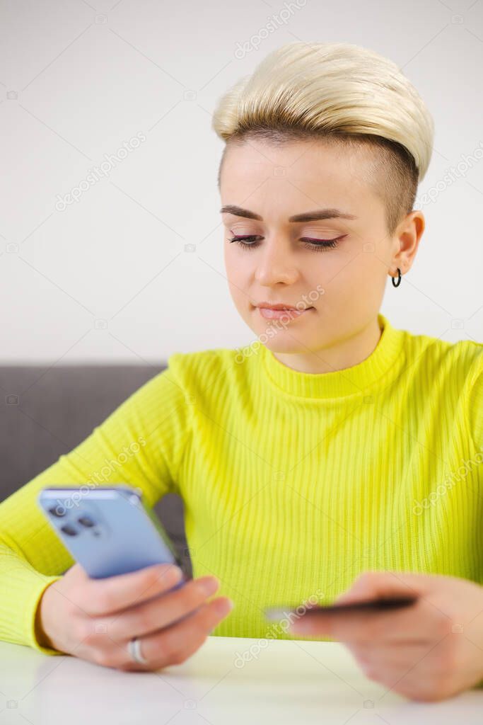 Young woman shopping online with credit card and modern mobile phone. Cool tom boy person with dyed short hair paying with bank card in app. Female transferring money with smartphone application