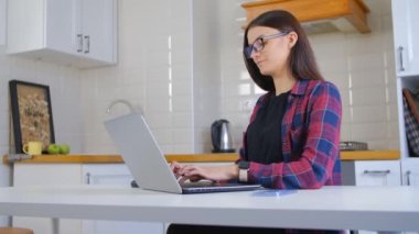 4K video 'da dizüstü bilgisayarda yazan genç girişimci kadın. Kilitleme sırasında ev mutfağında dizüstü bilgisayarda çalışan güzel beyaz bir kadın. Gözlük takan ve internetten uzak iş yapan inek.