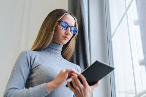 年轻的金发女人在家里的平板电脑上浏览互联网 漂亮的白色女性 戴着书呆子眼镜 身穿灰色高领汗衫 用现代小玩意进行在线交流 — 图库照片