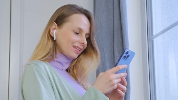 ワイヤレスヘッドフォンで音楽を聴き 携帯電話でメッセージを入力する幸せな白い女性 40代の陽気な白人女性がオンライン通信やエンターテイメントのために現代的な青のスマートフォンを使用して — ストック動画