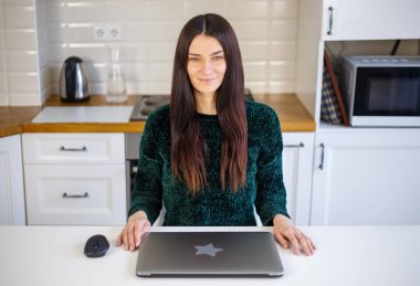 Dizüstü bilgisayarı olan güzel beyaz kadın. Ev mutfağında modern defter bilgisayarıyla poz veren uzun siyah saçlı mutlu, genç bir kadın. Serbest çalışan kişi neşe dolu bir gülümsemeyle kilit altında çalışmaya hazır.