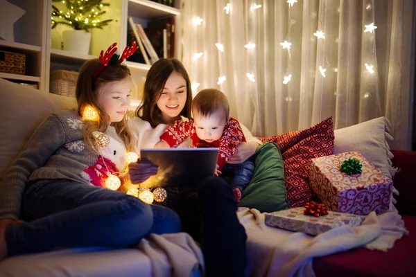 圣诞节的时候 两个女孩和一个男婴在温暖舒适的客厅里使用平板电脑 在圣诞节前夕有网上视频通话的家庭 冬夜与家人和孩子呆在家里 — 图库照片