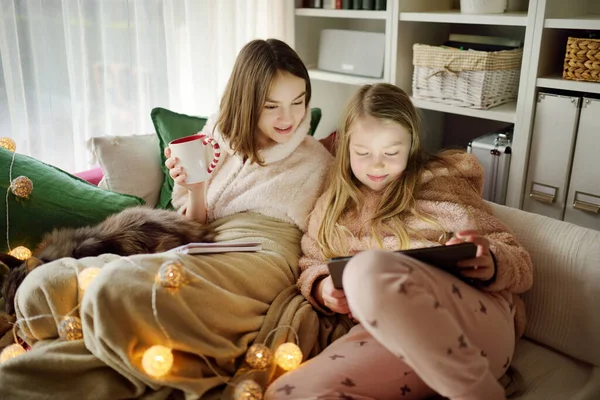 圣诞节的时候 两个妹妹依偎在一个舒适的客厅里的沙发上 寒假期间 可爱的孩子们在家里用平板电脑 孩子们躺在舒适的毯子里看书 在家里庆祝圣诞节 — 图库照片