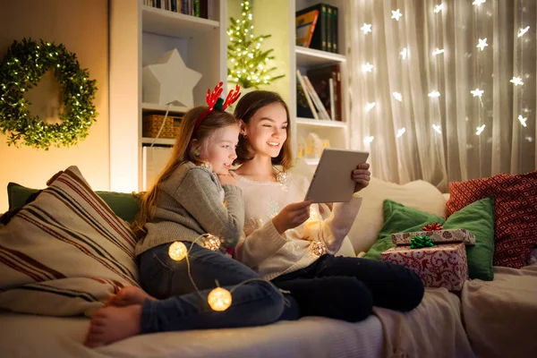 Two cute young sisters using a tablet pc at home in warm and cozy living room at Christmas. Family having online video call on Xmas eve. Winter evening at home with family and kids.