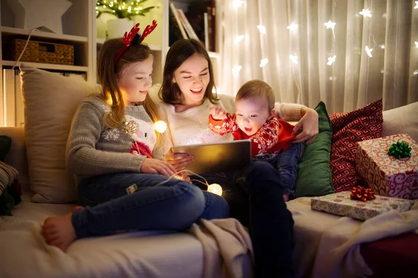 圣诞节的时候 两个女孩和一个男婴在温暖舒适的客厅里使用平板电脑 在圣诞节前夕有网上视频通话的家庭 冬夜与家人和孩子呆在家里 — 图库照片