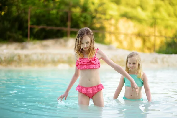 Two young sisters bathing in natural swimming pool in Bagno Vignoni, with thermal spring water and calcium carbonate deposits, which form white concretions and waterfall. Tuscany, Italy.