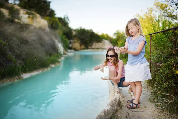 Two young sisters playing by natural swimming pool in Bagno Vignoni, with thermal spring water and calcium carbonate deposits, which form white concretions and waterfall. Tuscany, Italy.