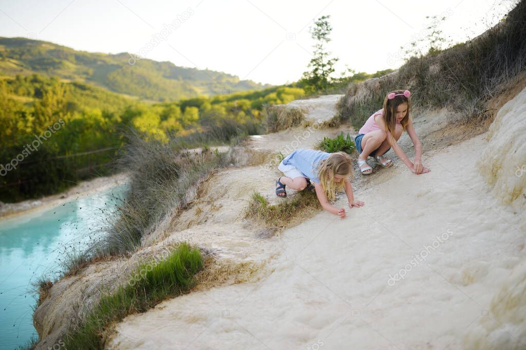 Two young sisters playing by natural swimming pool in Bagno Vignoni, with thermal spring water and calcium carbonate deposits, which form white concretions and waterfall. Tuscany, Italy.