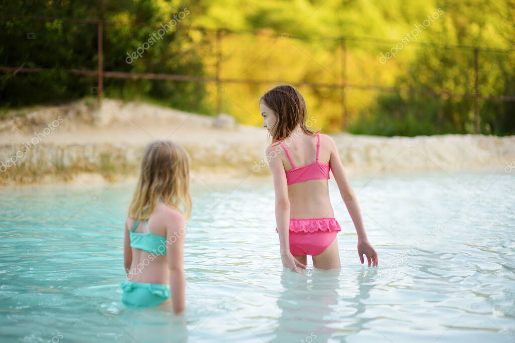 Two young sisters bathing in natural swimming pool in Bagno Vignoni, with thermal spring water and calcium carbonate deposits, which form white concretions and waterfall. Tuscany, Italy.