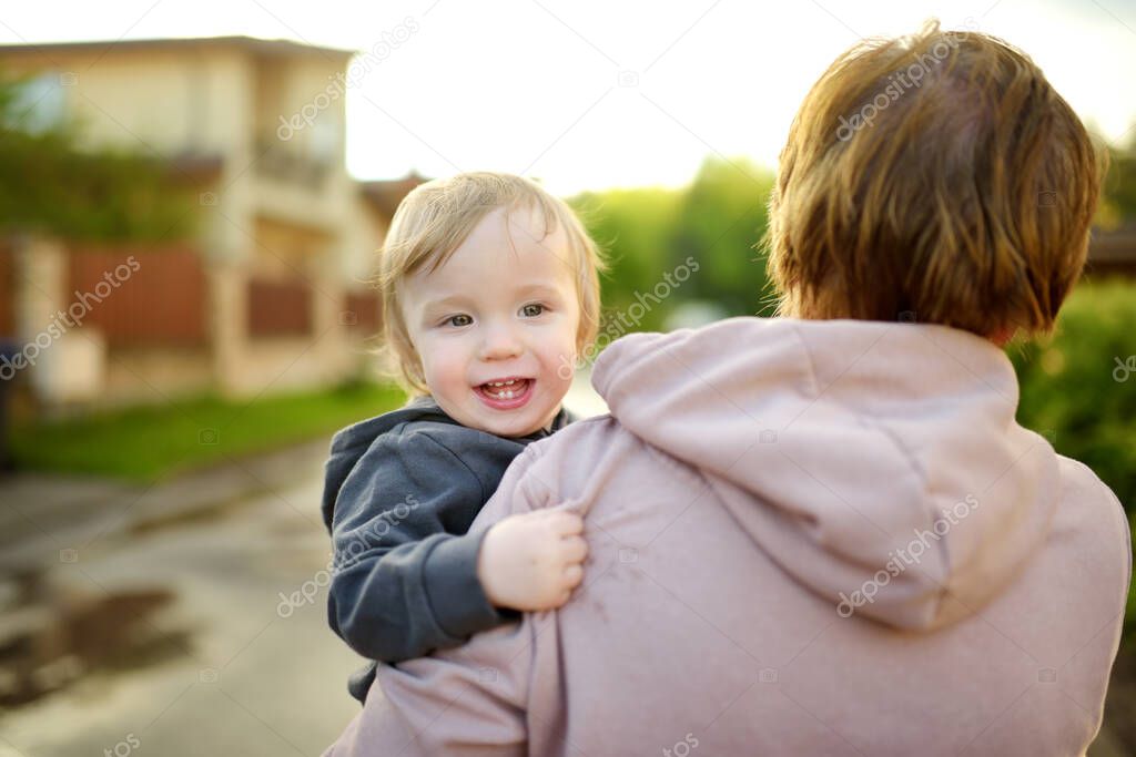 Cute funny toddler boy in his mothers arms. Mom and son having fun on sunny summer day in a park. Adorable son being held by his mommy.