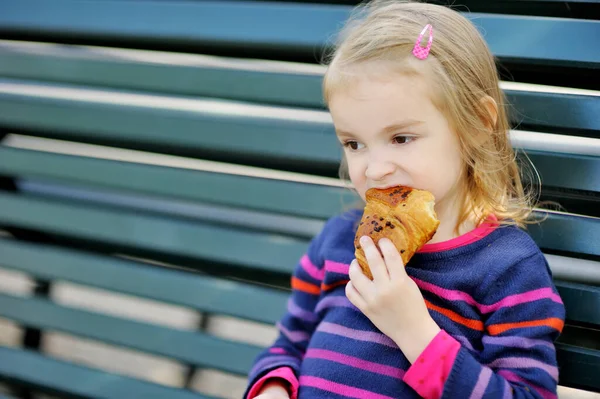 Adorable Little Girl Eating Fresh Baked Croissant While Sitting Bench — Stock Photo, Image