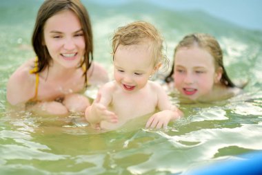 Şirin ablalar ve küçük erkek kardeşleri açık havuzda eğleniyorlar. Çocuk yüzmeyi öğreniyor. Çocuk su oyuncaklarıyla eğleniyor. Havuzda aile eğlencesi. Çocuklu aile için yaz etkinlikleri.