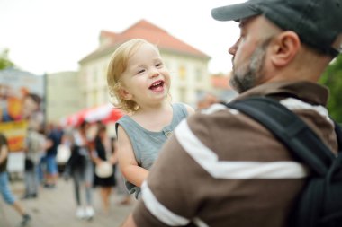 Babasının kollarında tatlı bir çocuk var. Baba ve oğul güneşli bir yaz gününde şehirde eğleniyorlar. Sevimli bebek babası tarafından tutuluyor..
