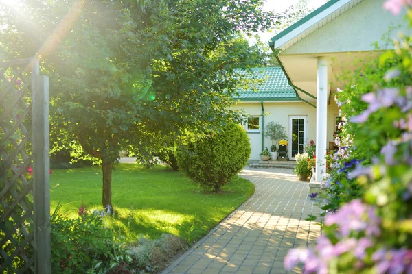 Ağaçlarla dolu güzel yeşil bir bahçe, dekoratif bitkiler ve arka planda büyük beyaz bir ev ile çiçek açan çiçekler. Arka bahçede güzel bir yaz günü.