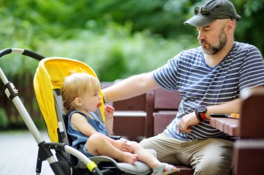 Baba, bebek arabasında oturan bebeğe yemek yediriyor. Baba ve oğul güneşli bir yaz gününde şehirde eğleniyorlar. Yazın dışarıda yemek yemek..