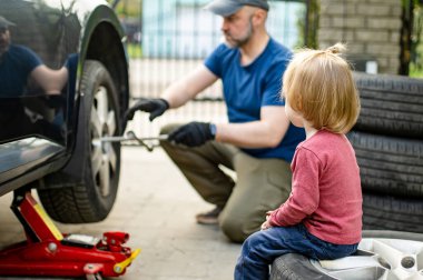 Tatlı çocuk, babasının arka bahçesinde araba tekerleklerini değiştirmesine yardım ediyor. Baba küçük oğluna alet kullanmayı öğretiyor. Küçük bir çocuğun aktif ebeveyni.