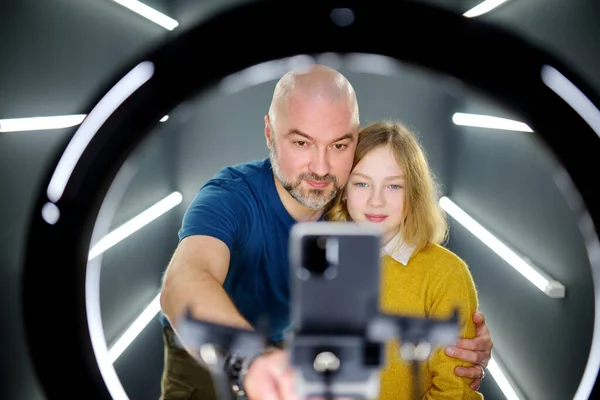 Father and his young daughter taking a selfie picture with mobile phone in light of the ring lamp. Trend, technology, beauty, fashion concept.
