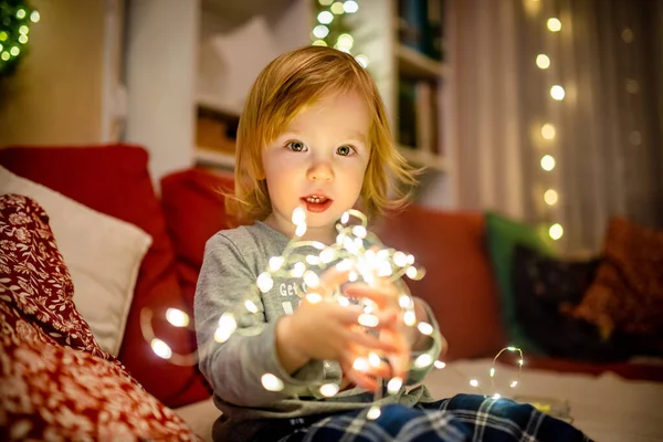 Happy little toddler boy playing with Chistmas lights in a cozy living room on Christmas eve. Celebrating Xmas at home. Winter evening with family and kids.