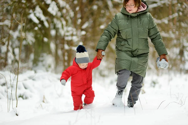 Cute toddler boy and his grandma having fun on a walk in snow covered pine forest on chilly winter day. Child exploring nature. Winter activities for small kids.