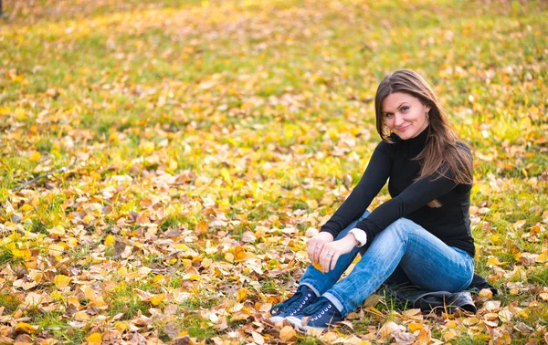 Giovane donna siede sulle foglie nel parco autunnale — Foto Stock