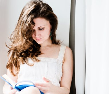 young caucasian brunette reading a book near a window at home