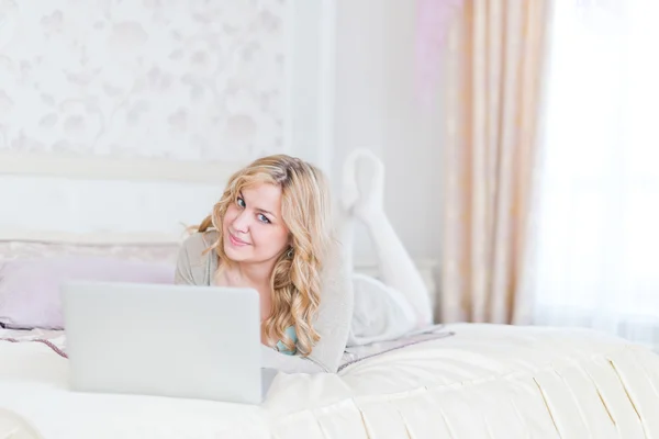 Feliz joven rubia acostada en su cama usando el ordenador portátil —  Fotos de Stock