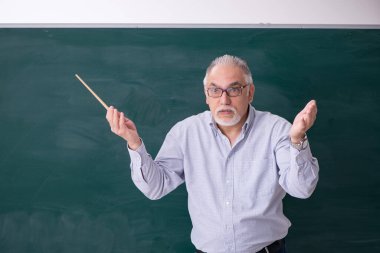 Yaşlı erkek öğretmen yeşil panonun önünde.