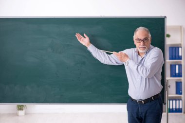 Yaşlı erkek öğretmen yeşil panonun önünde.