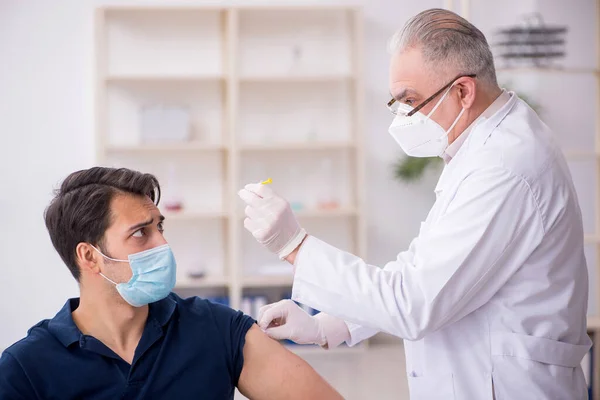 Jonge mannelijke patiënt bezoekt oude mannelijke arts in vaccinatiecentrum — Stockfoto