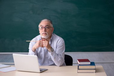 Yaşlı erkek öğretmen yeşil panonun önünde.