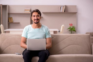 Evdeki bilgisayarla kanepede oturan genç adam.