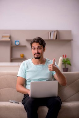 Evdeki bilgisayarla kanepede oturan genç adam.