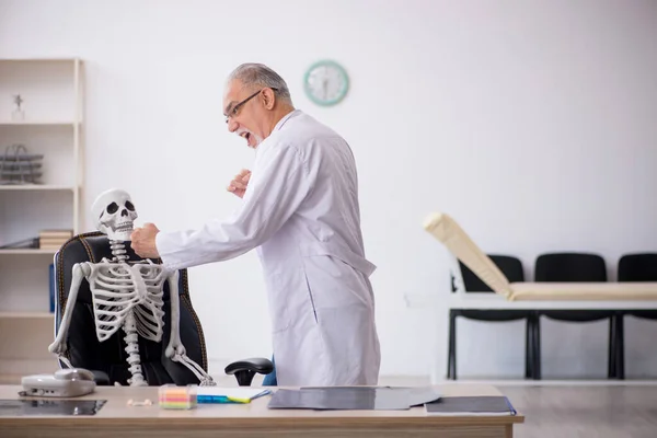 Vecchio medico maschio con scheletro in clinica — Foto Stock