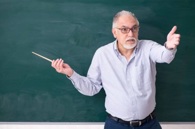 Yaşlı erkek öğretmen yeşil panonun önünde.