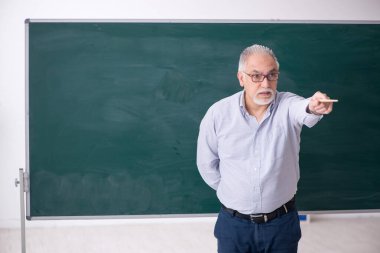 Yaşlı erkek öğretmen yeşil panonun önünde.