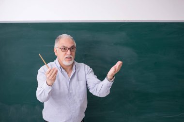 Yaşlı erkek öğretmen yeşil panonun önünde.