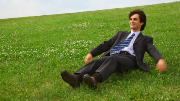 Красивый мужчина в костюме ложится на зеленую траву и поднимает ноги — стоковое видео