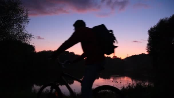 Man with rucksack on bicycle looks at sunset lake — Stock Video