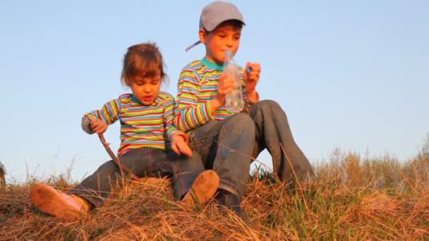 Хлопчик і дівчинка сидять на траві. Він п'є воду, вона щось говорить. — стокове відео