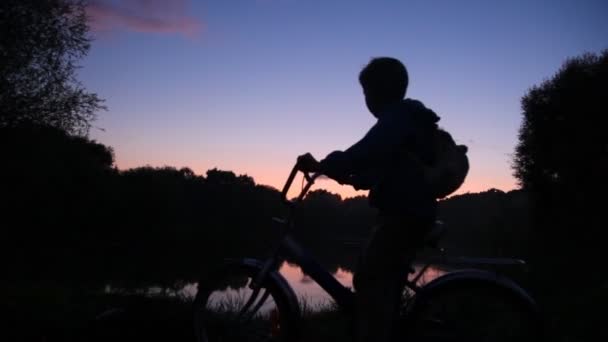 Boy with rucksack on bicycle looks at sunset lake — Stock Video