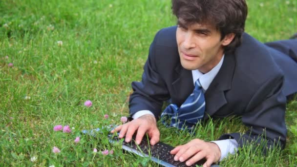 Красивый человек, печатающий на клавиатуре, лежит на зеленой траве, вид спереди — стоковое видео