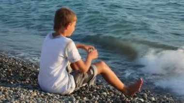 genç çocuk çakıl plajı üzerinde oturur ve denizde, arka tarafı görünümü görünüyor