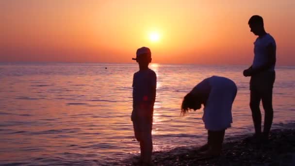 Family throwing pebbles to water, sunset sea in background — Stock Video