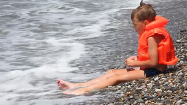 Menino vestido com jaqueta inflável senta-se na praia de seixos com mar surf — Vídeo de Stock
